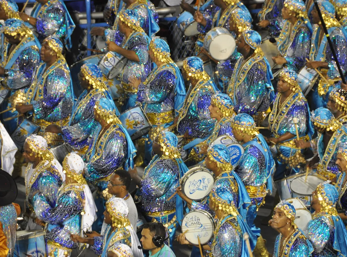 A samba school sweeps down the Sambadrome runway at night in towering feathered costumes lit by floodlights