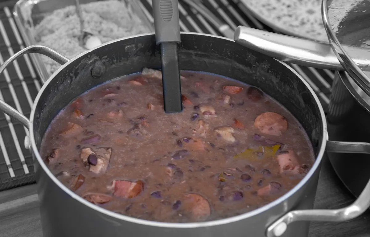 A steaming clay pot of feijoada surrounded by rice, sliced oranges, collard greens and farofa on a wooden table