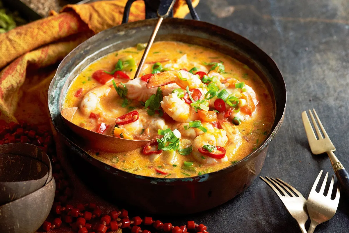 An orange-red moqueca bubbling in a clay pot, dotted with shrimp, peppers and coriander